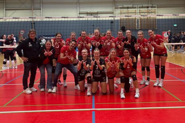 Die Spielerinnen und das Trainerteam der BayerVolleys posieren nach dem WVV-Pokalsieg in roten Trikots auf dem Volleyballfeld.